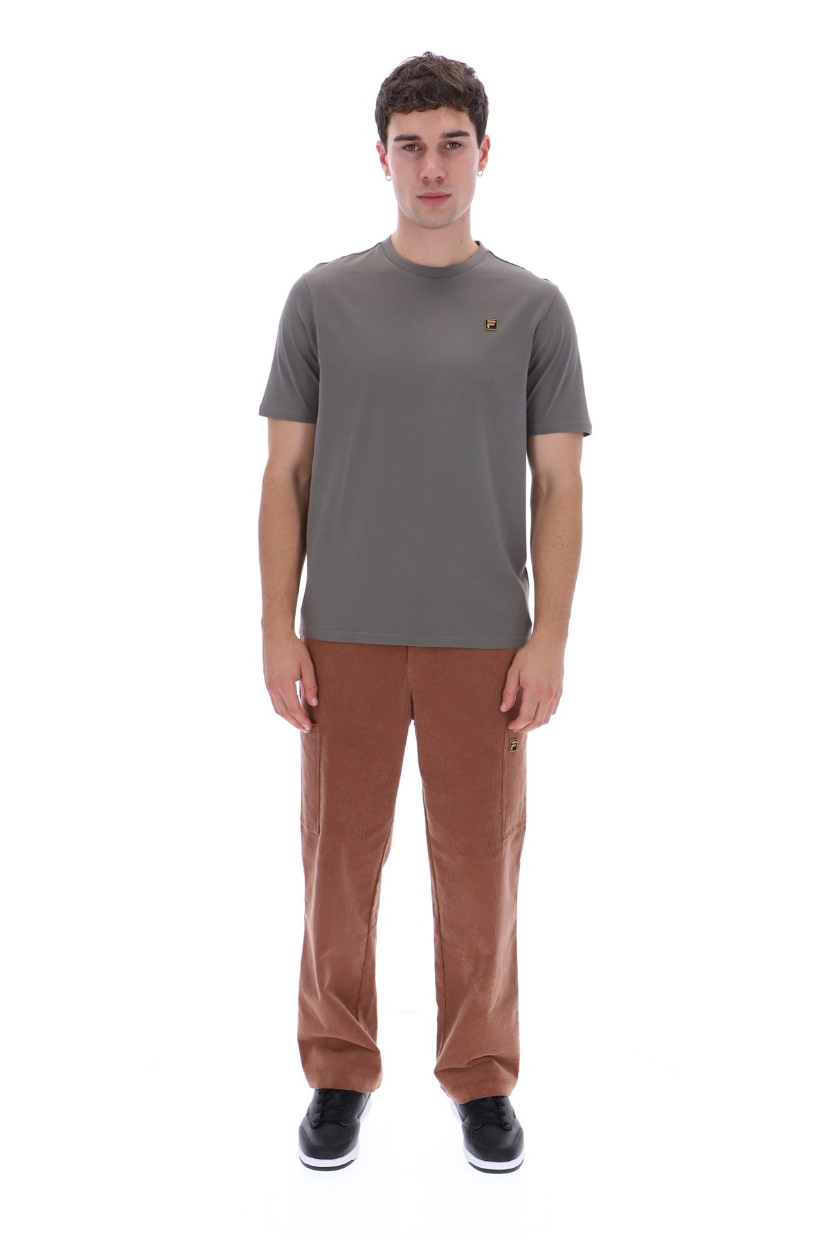 A man modeling a short-sleeve crewneck t-shirt in dark gray with a small embroidered logo on the chest, paired with brown pants and black sneakers, against a plain white background.