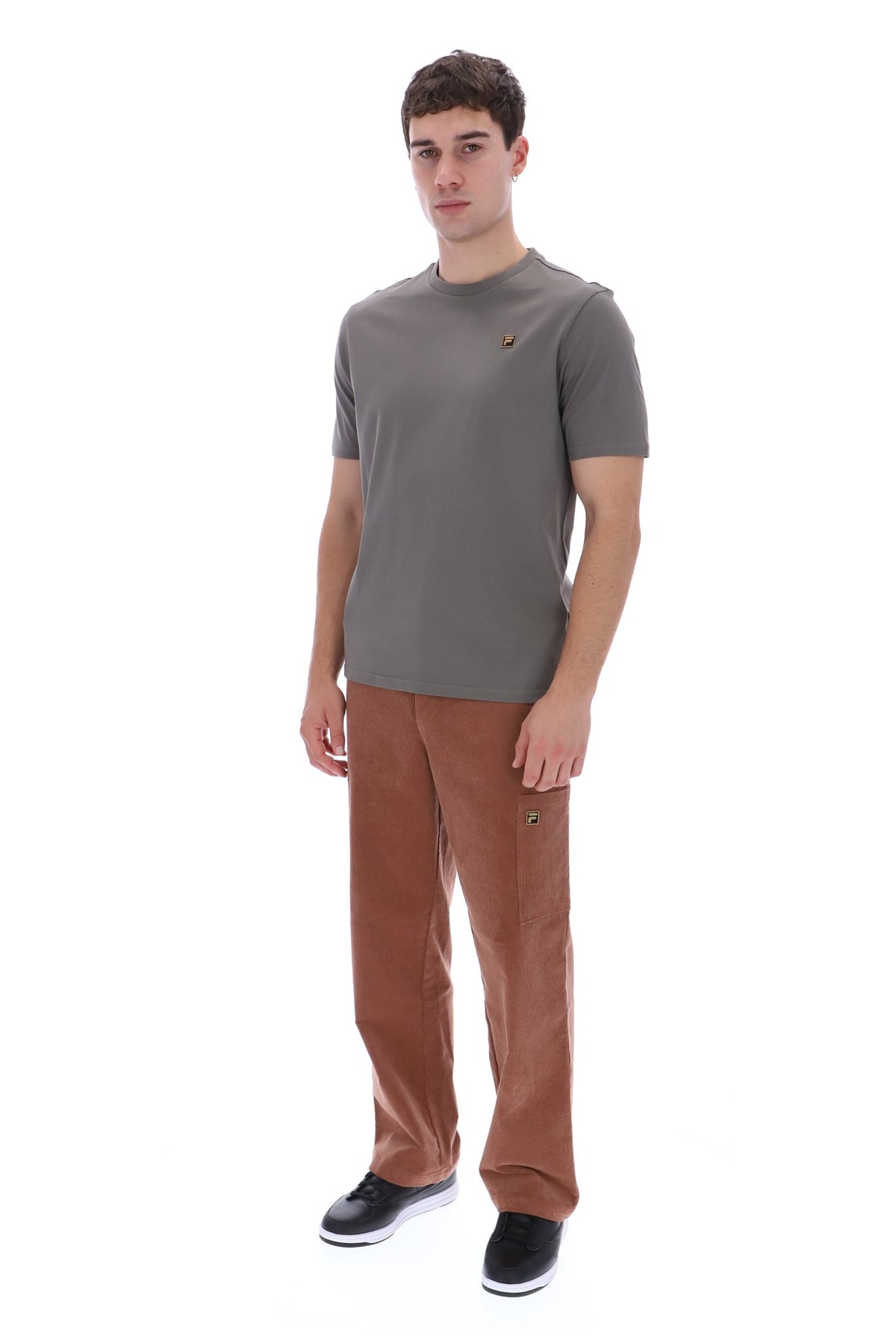 A man modeling a short-sleeve crewneck t-shirt in dark gray and brown corduroy trousers, standing against a solid white background.