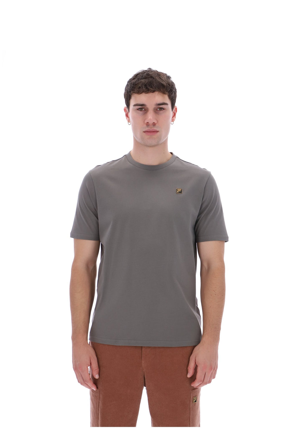 A man wearing a short-sleeve crewneck t-shirt in a muted gray-brown color, featuring a small embroidered logo on the left chest, paired with brown pants, against a solid white background.