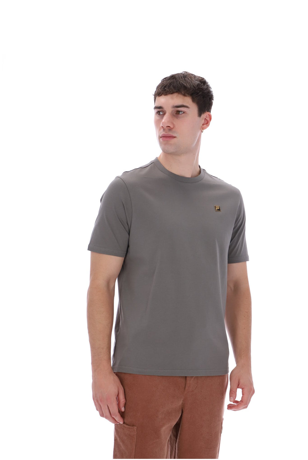 A man wearing a short-sleeve crewneck t-shirt in a muted gray-brown color, featuring a small embroidered bird logo on the left chest, paired with brown pants, against a solid white background.