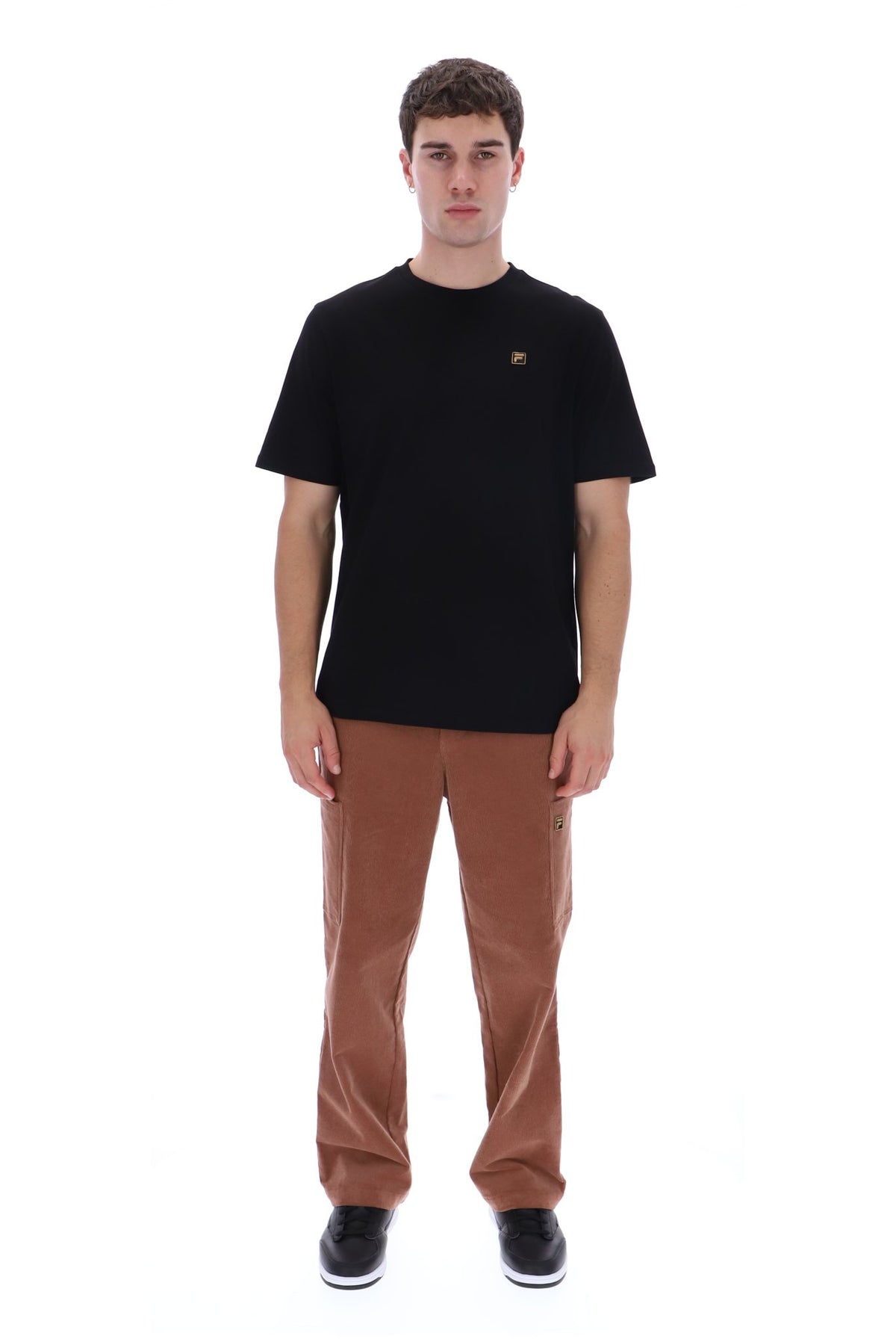 A man modeling a black short-sleeve crewneck t-shirt with a small embroidered logo on the left chest, paired with brown corduroy pants and black sneakers, against a plain white background.