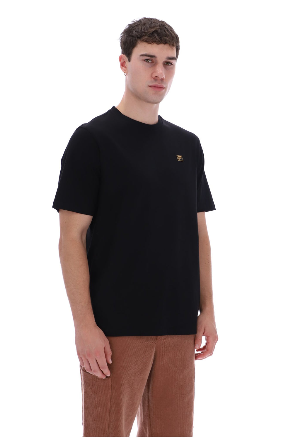 A man wearing a black short-sleeve crewneck t-shirt with a small gold embroidered logo on the left chest, paired with brown corduroy pants, against a plain white background.