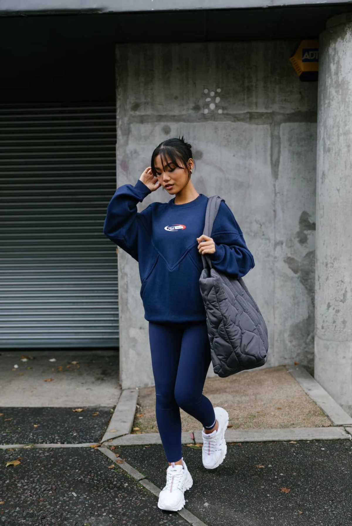 A person wearing high-waisted navy blue leggings with the FILA x TALA brand logo on the upper left thigh. They are also wearing a matching navy long-sleeve top and white sneakers, carrying a sports bag, and standing against an urban backdrop.