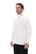 A man wearing a white long-sleeve mock neck turtleneck sweater with a small Fila logo on the collar, paired with black pants, photographed against a solid white background.