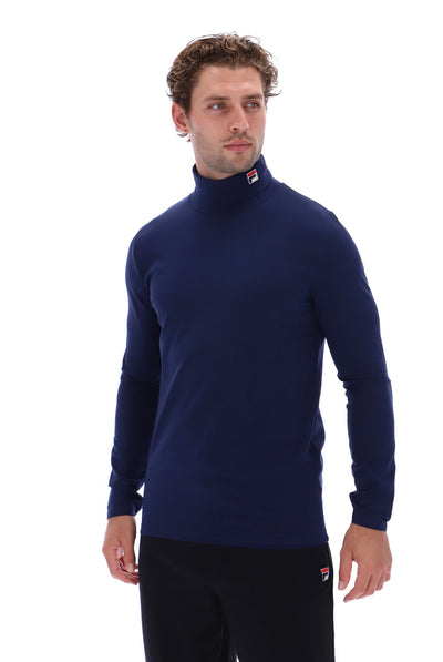 A man wearing a navy blue long-sleeve mock neck sweater and black pants, standing against a solid white background. The sweater features a small red and white Fila logo on the collar.