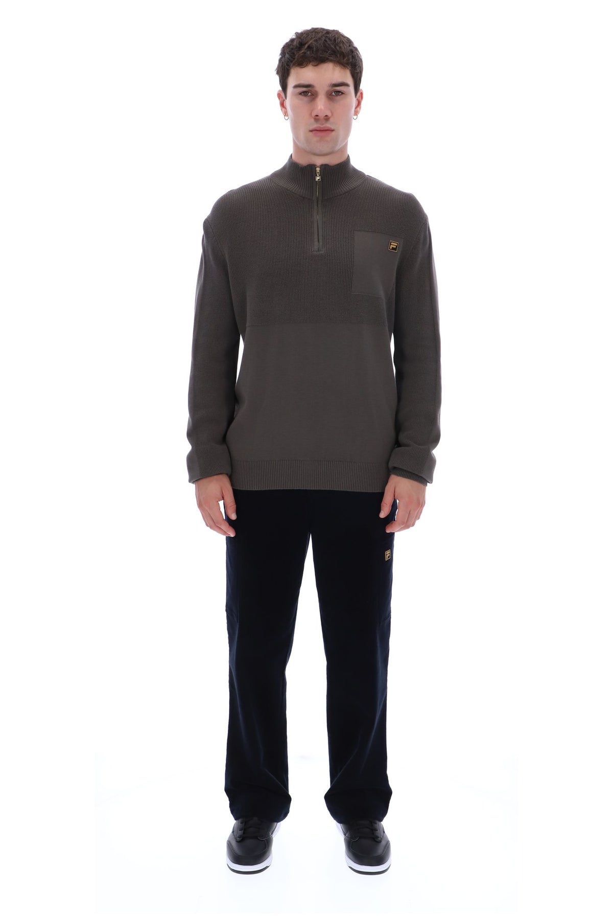 A man modeling a dark brown quarter-zip pullover sweater with a chest pocket and a small orange logo, paired with black pants and white sneakers, against a plain white background.
