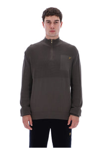 A man wearing a dark brown quarter-zip pullover sweater with a chest pocket and a small gold embroidered logo, paired with black pants, against a plain white background.