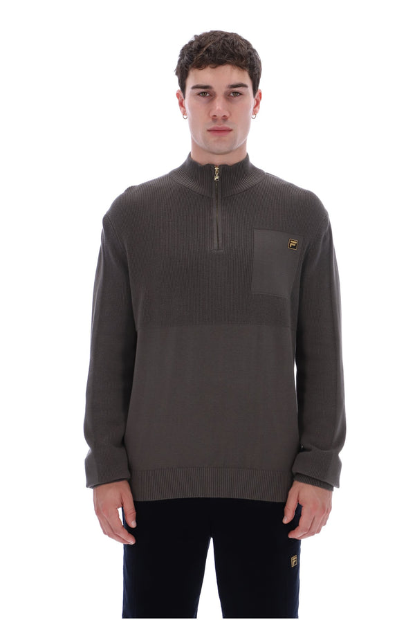 A man wearing a dark brown quarter-zip pullover sweater with a chest pocket and a small gold embroidered logo, paired with black pants, against a plain white background.