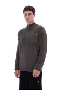 A man wearing a dark brown quarter-zip pullover sweater with a chest pocket and a small yellow logo, paired with dark pants, against a plain white background.