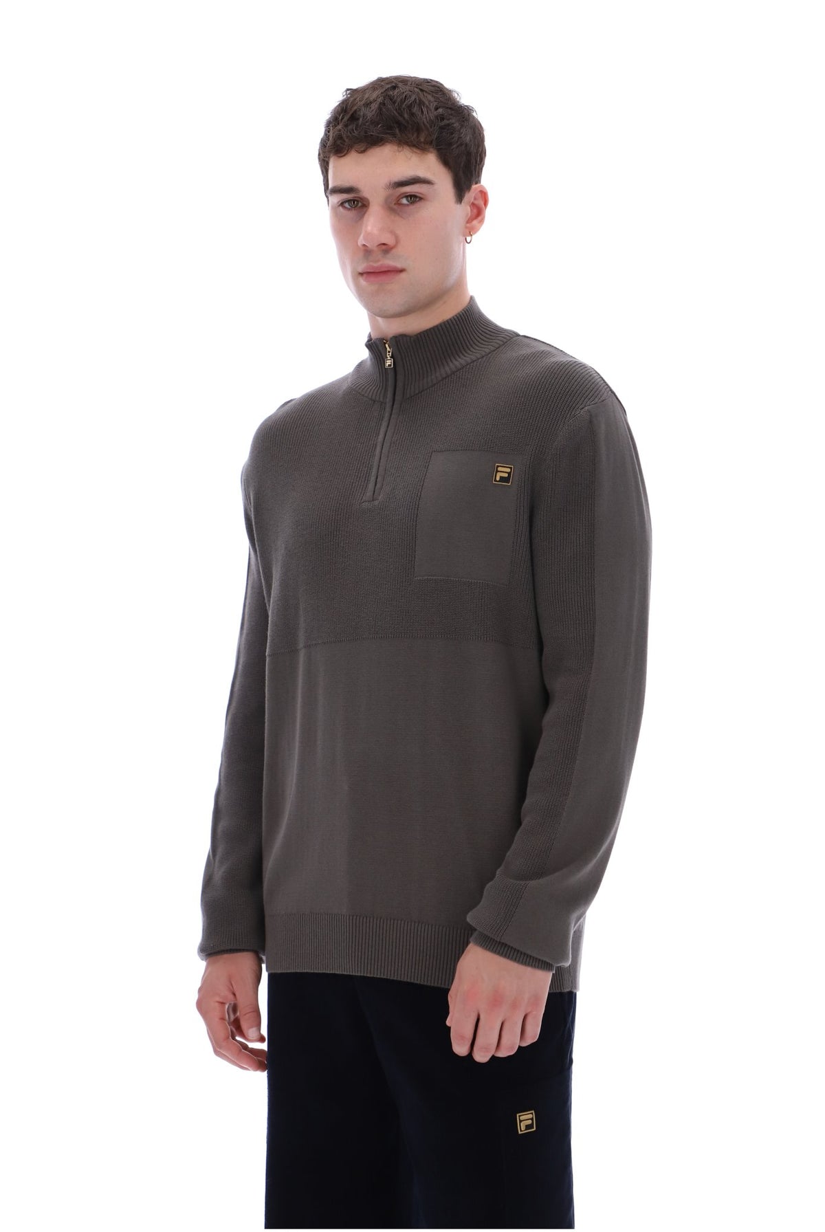 A man wearing a dark brown quarter-zip pullover sweater with a chest pocket and a small yellow logo, paired with dark pants, against a plain white background.