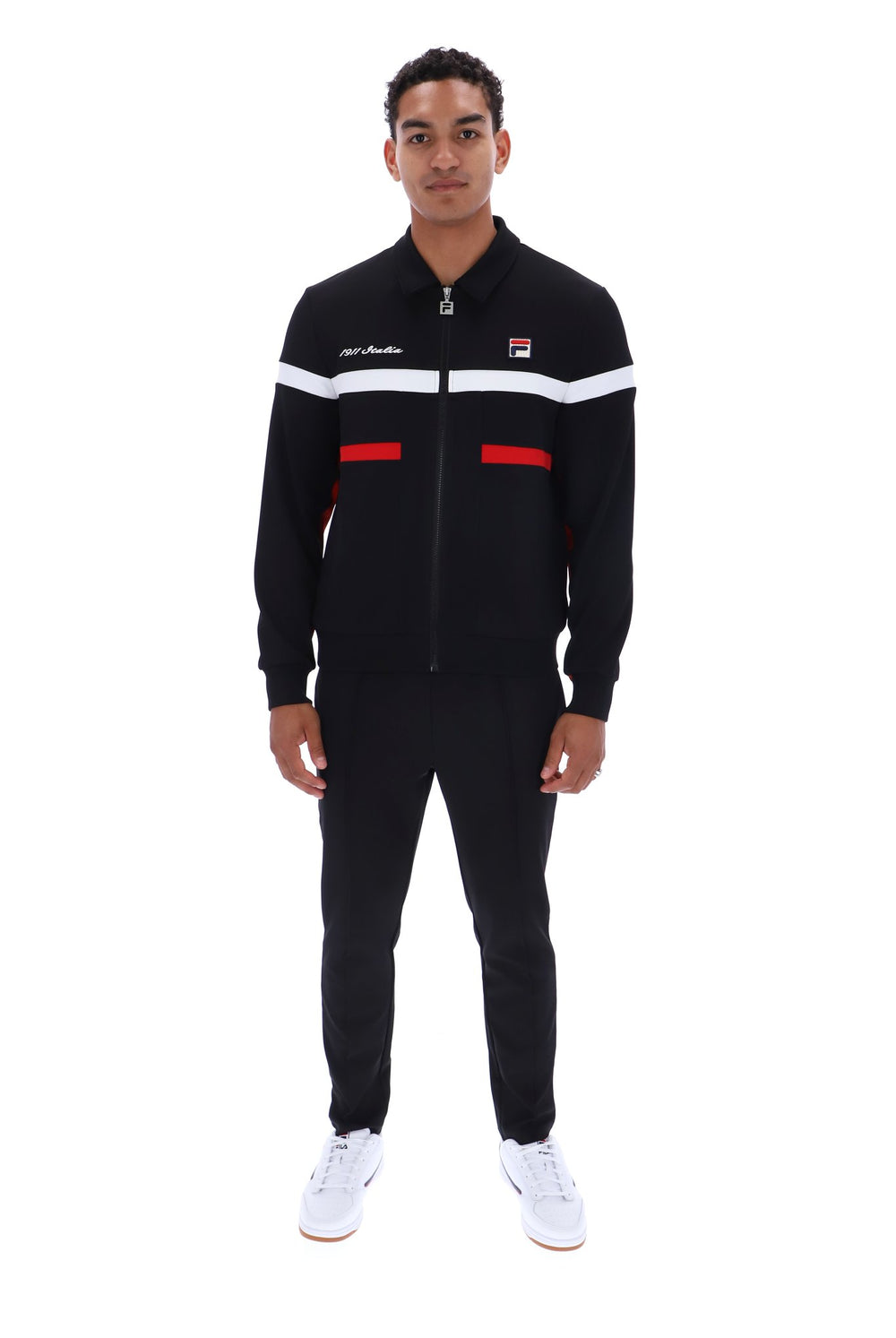 A man modeling a black FILA track suit with white and red stripe details on the chest and waist, paired with white sneakers, shot against a plain white background.