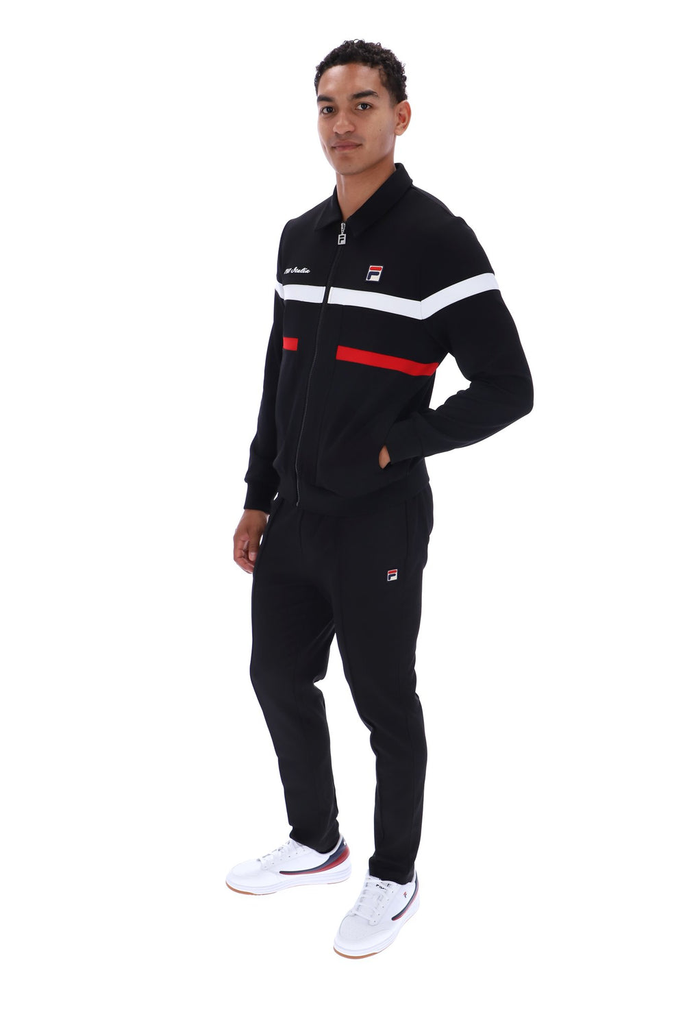 A man modeling a black FILA tracksuit with white and red stripe details, paired with white sneakers, against a plain white background.