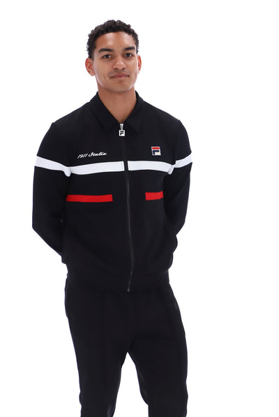 A man wearing a black Fila zip-up track jacket with white and red stripe accents, paired with black track pants, against a plain white background.