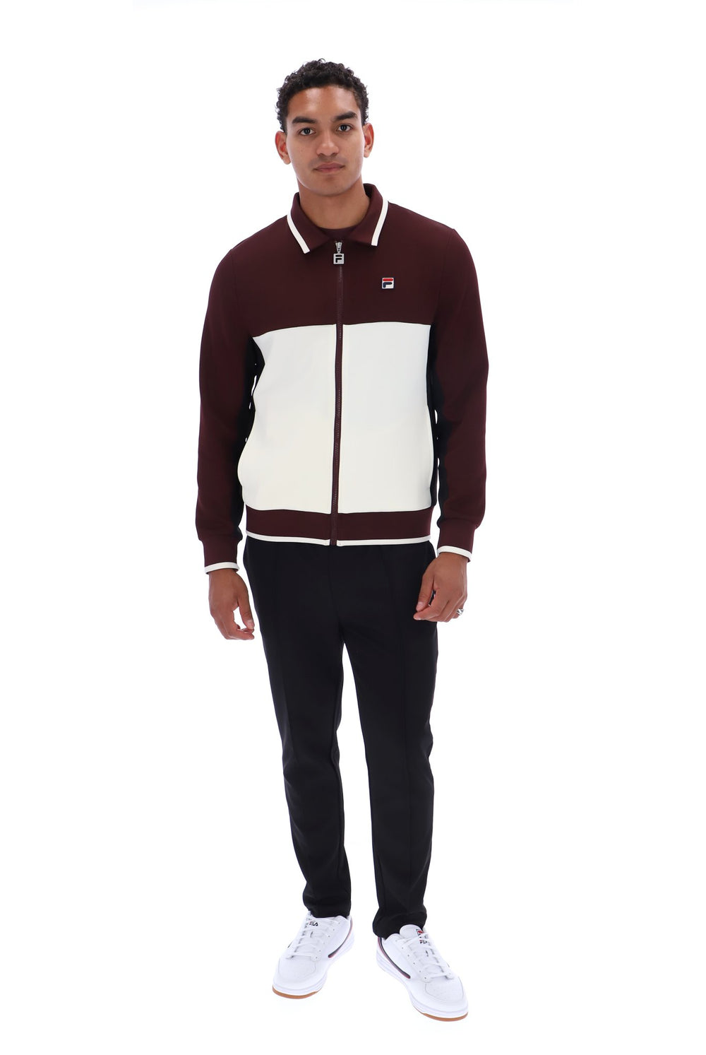 A man modeling a color-block zip-up jacket with dark maroon sleeves and front, and an off-white chest panel, paired with black pants and white sneakers, on a solid white background.