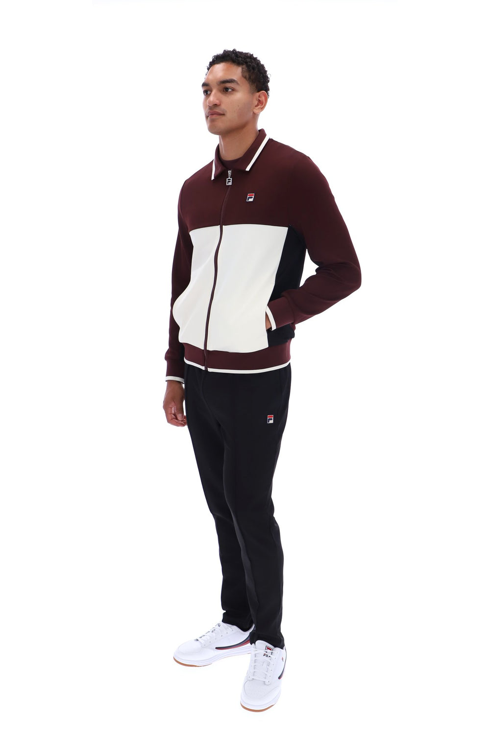 A man modeling a two-piece tracksuit from Filson, featuring a color-blocked zip-up jacket in burgundy, cream, and black, paired with black joggers and white sneakers, on a solid white background.