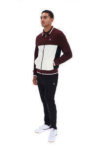 A man modeling a two-piece tracksuit from Filson, featuring a color-blocked zip-up jacket in burgundy, cream, and black, paired with black joggers and white sneakers, on a solid white background.