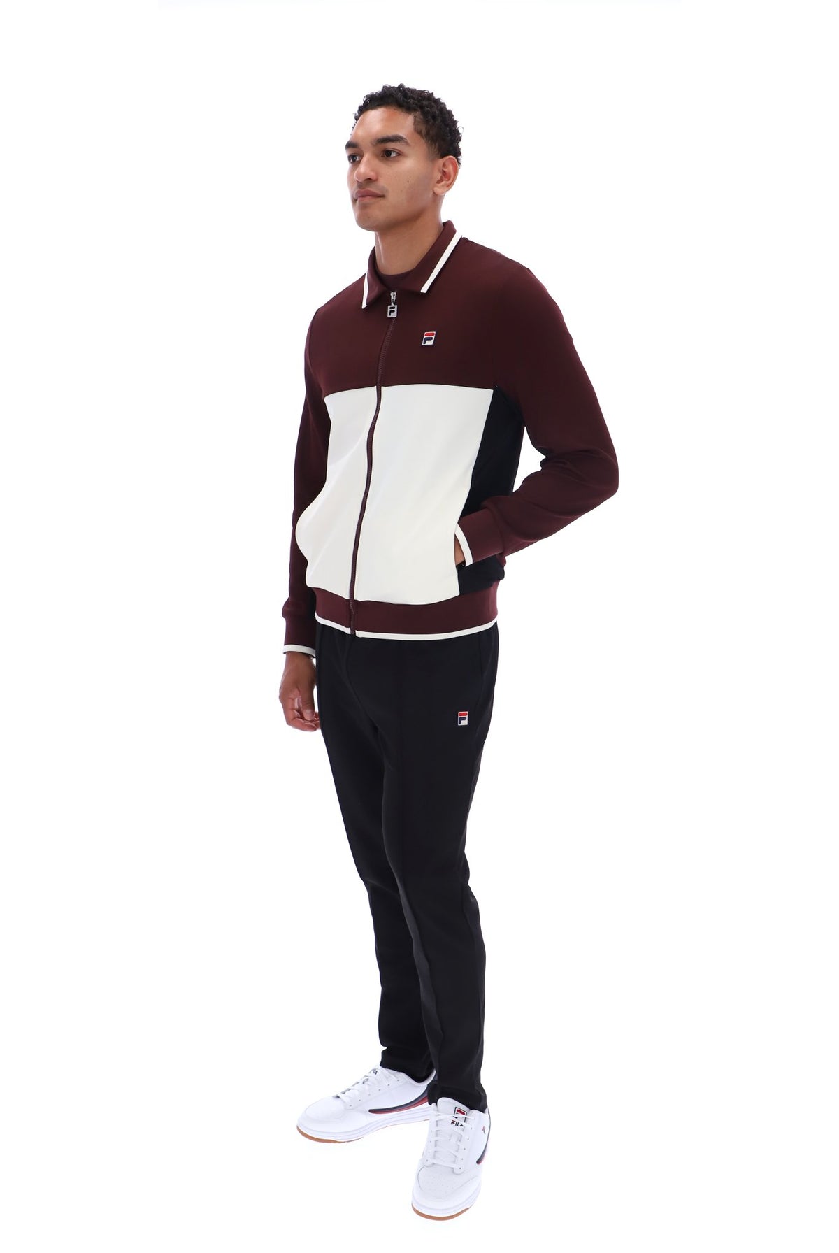 A man modeling a two-piece tracksuit from Filson, featuring a color-blocked zip-up jacket in burgundy, cream, and black, paired with black joggers and white sneakers, on a solid white background.