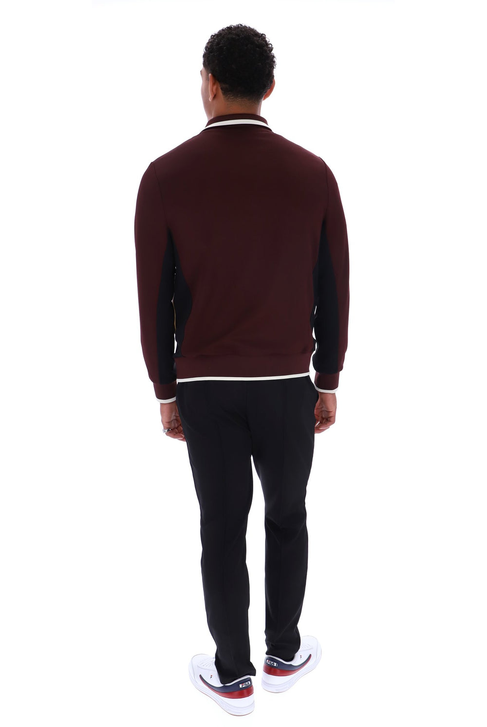 A man viewed from behind wearing a burgundy and black long-sleeve knit sweater with white trim, black pants, and white sneakers, shot against a plain white background.