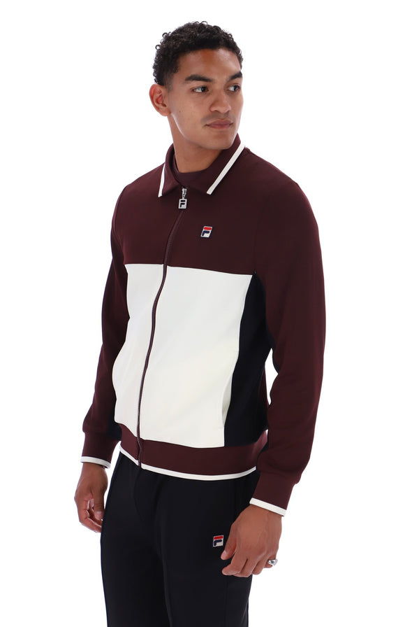 A man wearing a Fila colorblock zip-up track jacket in maroon, white, and black, paired with black track pants, against a plain white background.