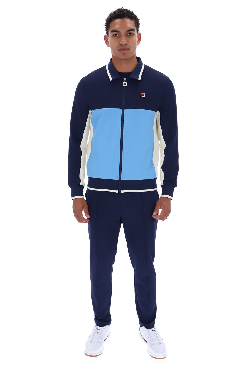 A man modeling a navy blue, light blue, and white color-blocked track suit with white sneakers against a plain white background.