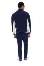 A man viewed from the back wearing a navy blue knit tracksuit with white contrast panels and white sneakers, set against a plain white background.