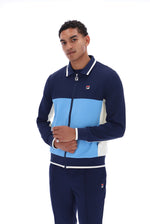 A man modeling a navy blue, light blue, and white color-blocked zip-up track jacket and matching navy pants by Fila, set against a plain white background.