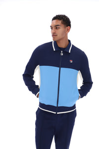 A man wearing a color-blocked navy blue and light blue track jacket with white trim, featuring the Fila logo on the chest, against a solid white background.