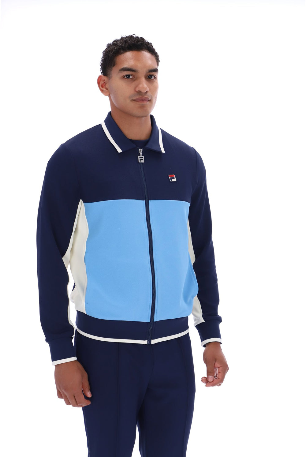 A man wearing a Fila colorblock zip-up track jacket and matching navy pants against a white studio background.