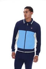 A man wearing a navy blue, light blue, and white color-blocked zip-up track jacket and matching navy pants, featuring the Fila logo on the chest and sleeve.