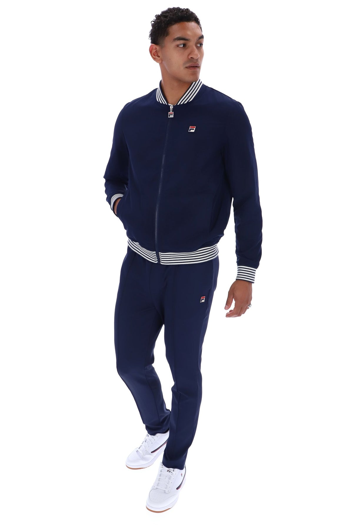 A man modeling a navy blue tracksuit with white and navy striped ribbed trim, featuring a zip-up jacket and matching sweatpants, on a solid white background.