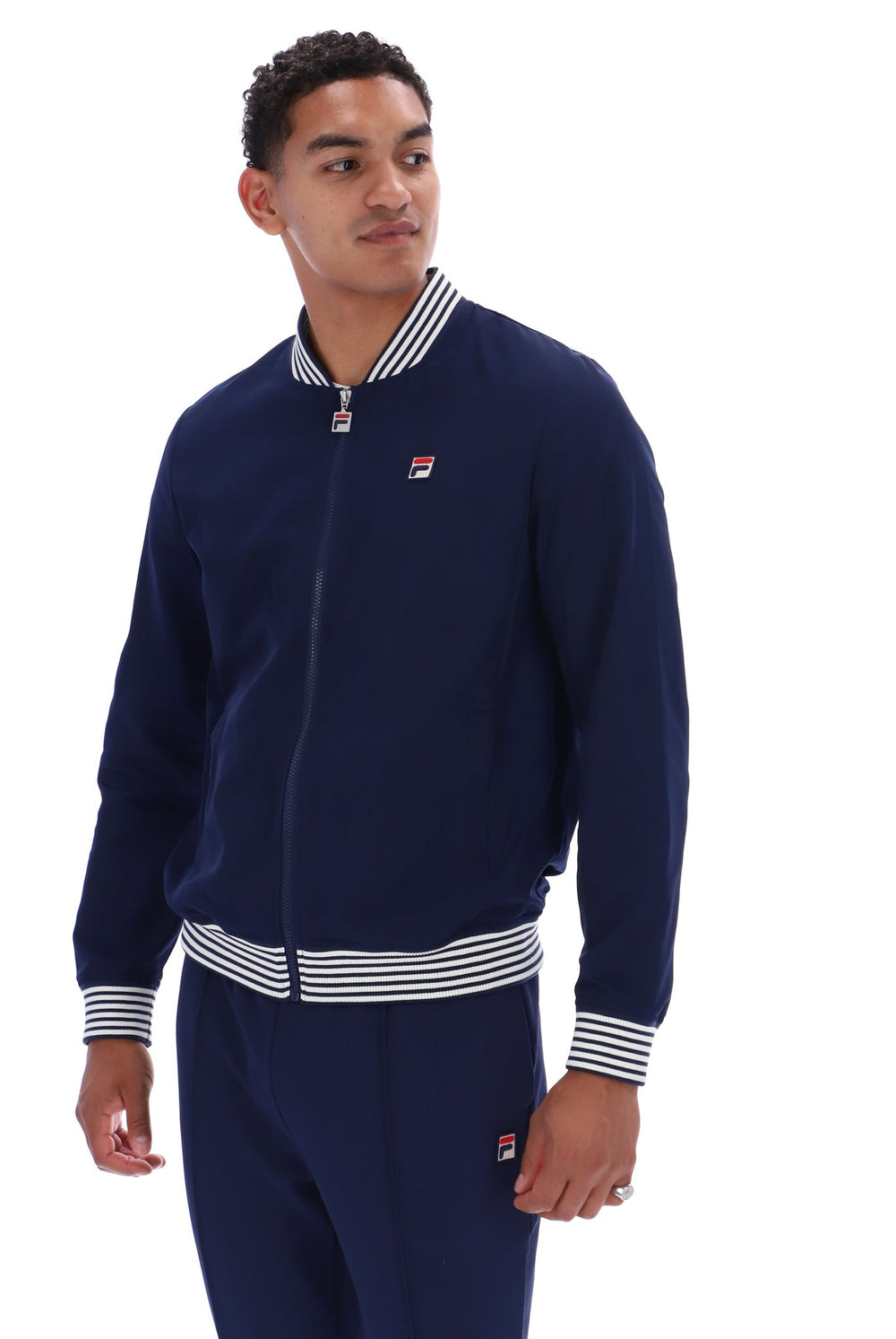 A man wearing a navy blue Fila zip-up track jacket with white striped ribbed collar, cuffs, and hem, paired with matching navy pants, against a plain white background.