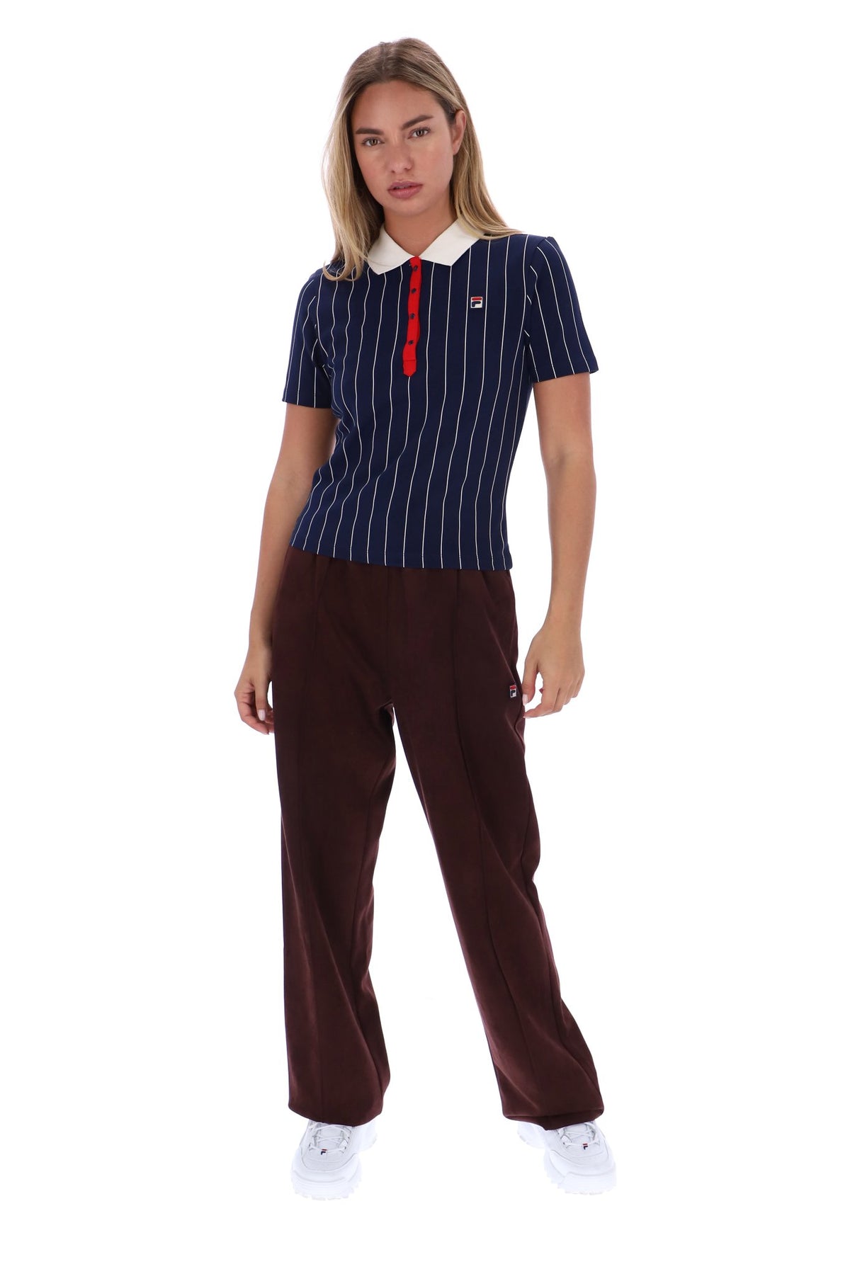 A woman modeling a short-sleeve polo shirt with navy blue and white vertical pinstripes, a white collar, and a red placket, paired with dark brown trousers and white sneakers, against a plain white background.