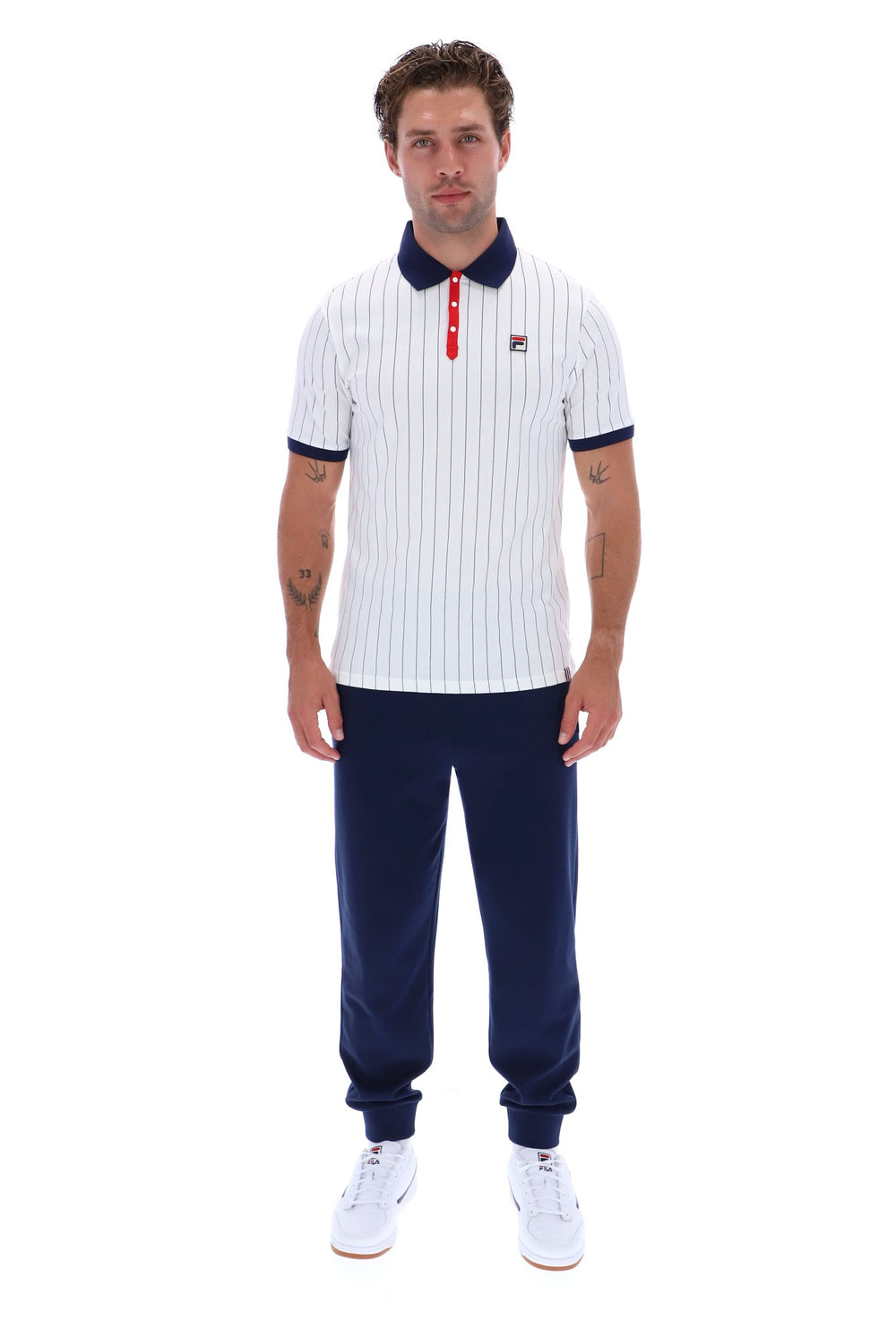 A man modeling a white short-sleeve polo shirt with navy blue pinstripes, a navy collar, and a red button placket, paired with navy pants and white sneakers, against a plain white background.