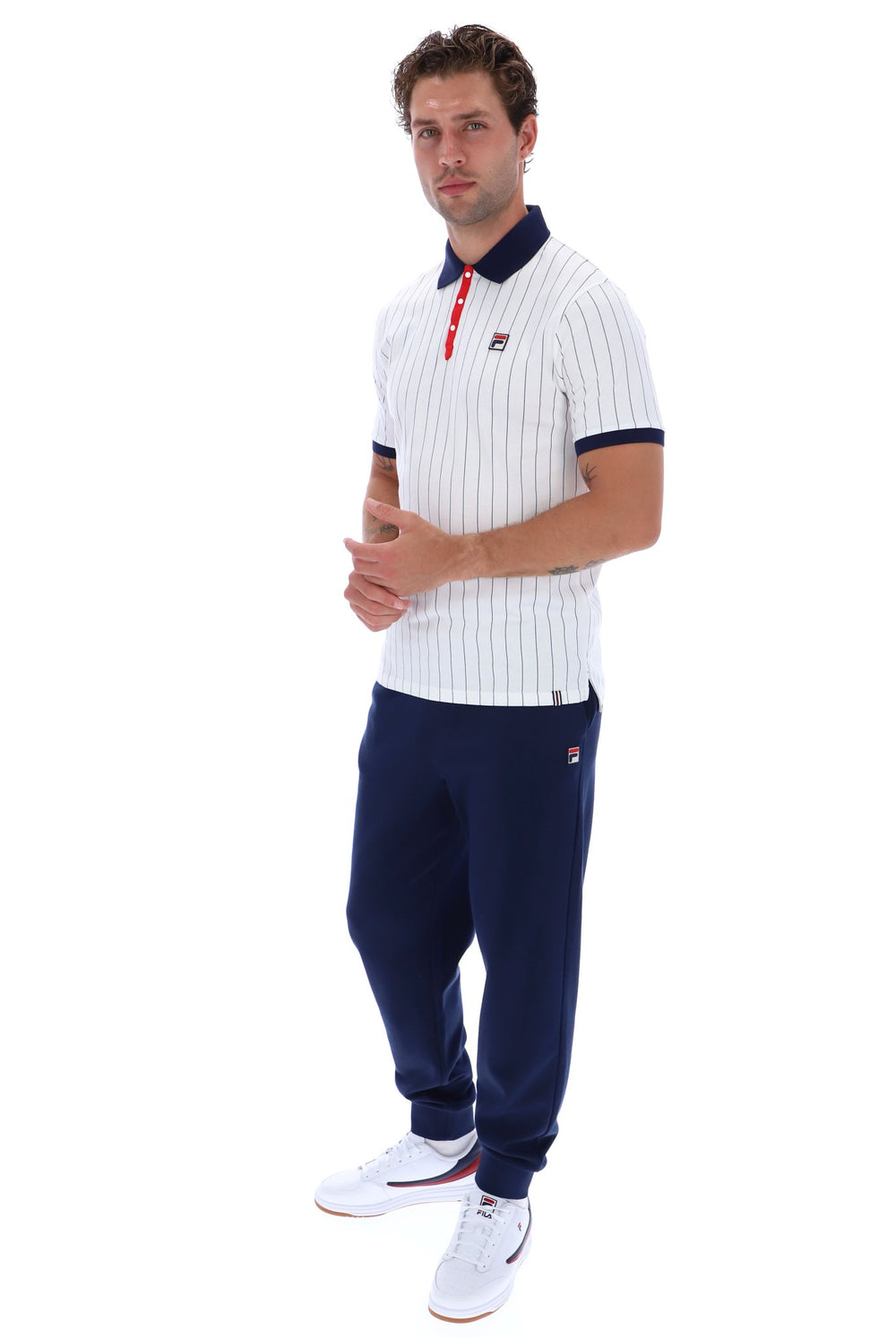 A man modeling a white short-sleeve polo shirt with thin navy vertical pinstripes, a navy collar, and a small red-and-white logo on the chest, paired with navy jogger pants and white sneakers, against a plain white background.