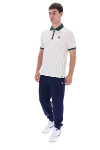 A man modeling a white short-sleeve polo shirt with thin green stripes, a green collar, and a green trim on the collar and sleeve cuffs, paired with navy blue pants and white sneakers. The polo shirt features a small red and white Fila logo on the left chest.