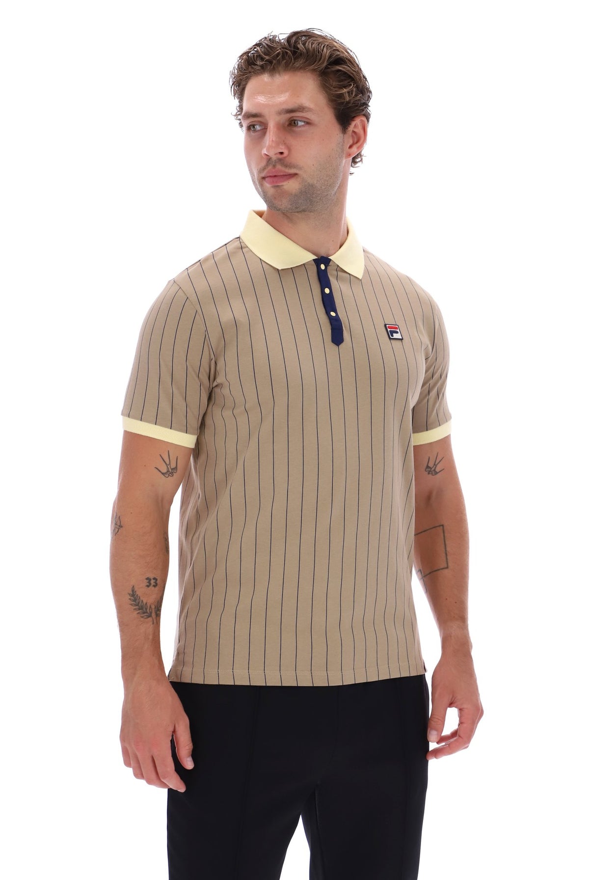 A man wearing a beige short-sleeve polo shirt with thin vertical navy stripes, a light yellow collar and sleeve cuffs, and a small red, white, and blue FILA logo on the left chest, paired with black pants against a white background.