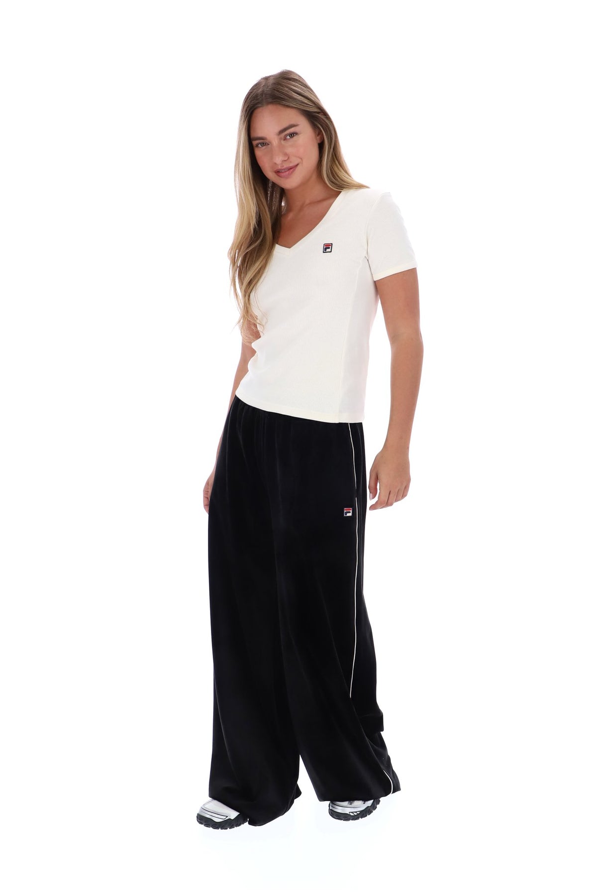 A woman modeling a white short-sleeve V-neck t-shirt and black wide-leg sweatpants with a small red, white, and blue FILA logo on the chest and thigh.