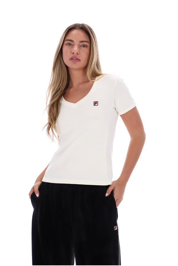 A woman modeling a white short-sleeve V-neck T-shirt with a small red, white, and blue FILA logo on the left chest, paired with black pants, against a solid white background.