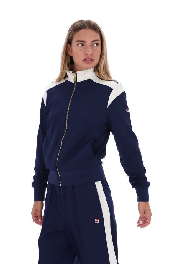 A woman modeling a navy blue tracksuit with white shoulder panels and white side stripes, featuring a gold zipper and a small red and white logo patch.