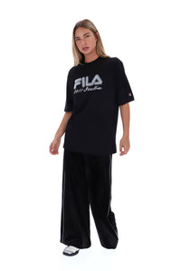 A woman modeling an oversized black FILA t-shirt with white logo text, paired with black wide-leg pants with white side stripes, on a solid white background.