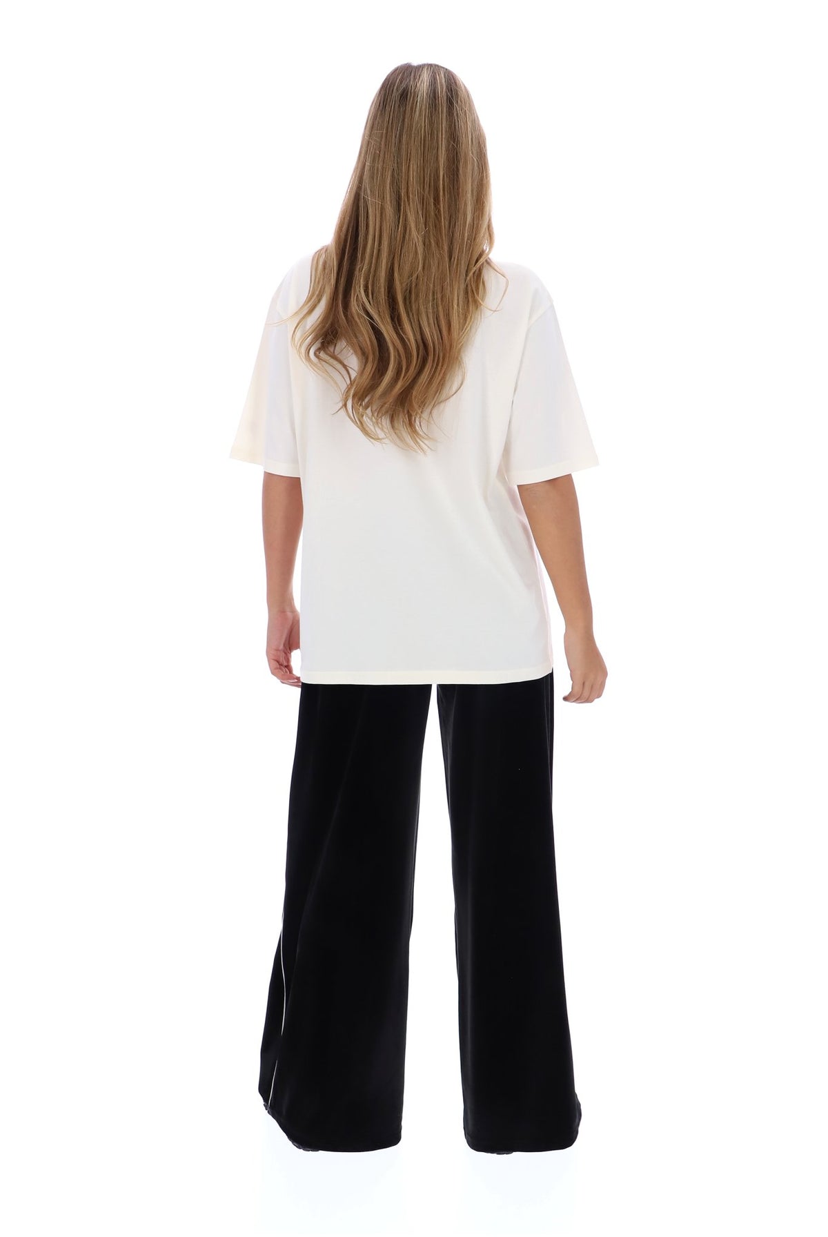 A woman viewed from the back wearing an oversized off-white short-sleeve top and black wide-leg pants, standing against a plain white studio background.