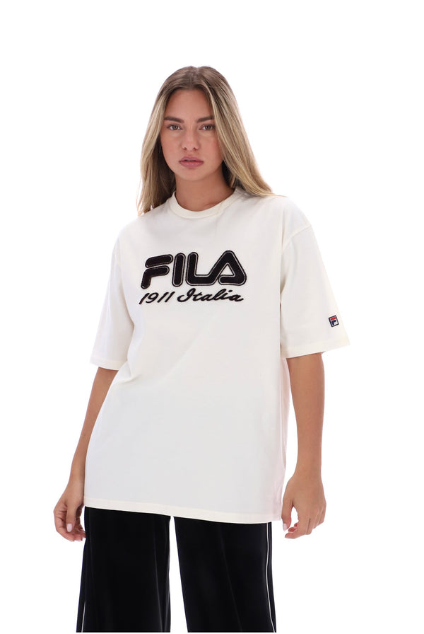 A woman modeling a white FILA t-shirt with a black graphic logo on the chest, paired with black pants, against a plain white background.