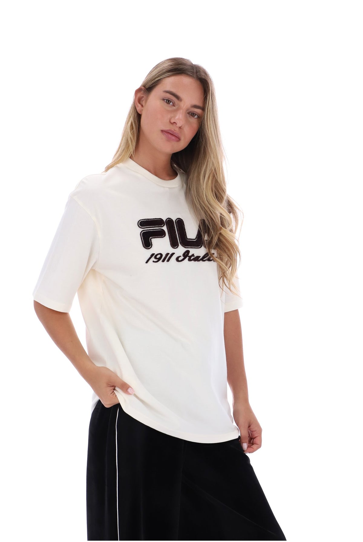 A woman modeling a white short-sleeve t-shirt with a black FILA 1911 Studio logo on the chest, paired with black pants, against a solid white background.