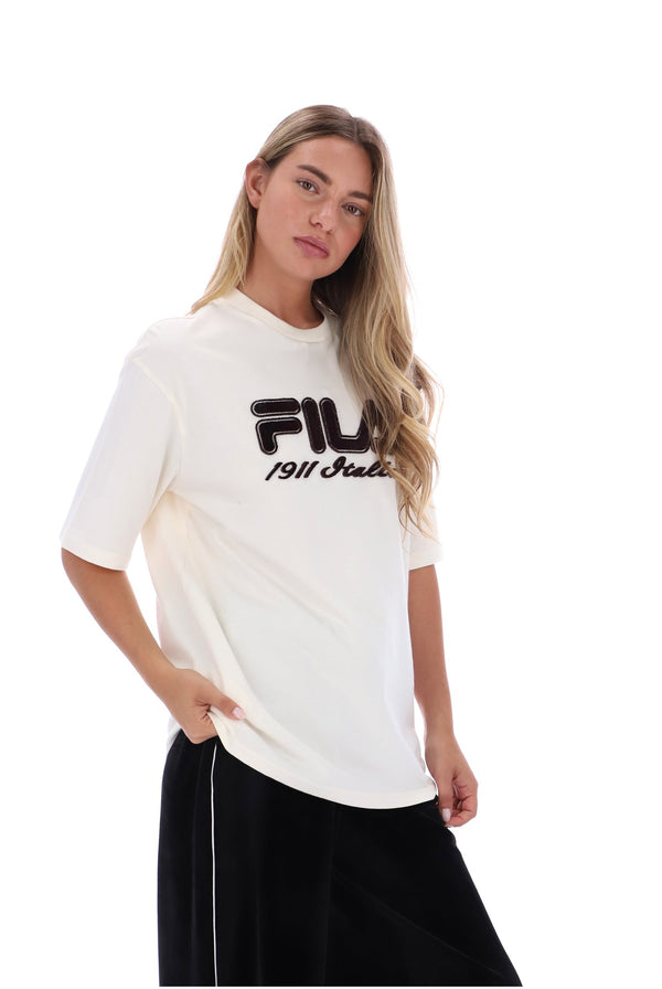 A woman modeling a white short-sleeve t-shirt with a black FILA 1911 Studio logo on the chest, paired with black pants, against a solid white background.