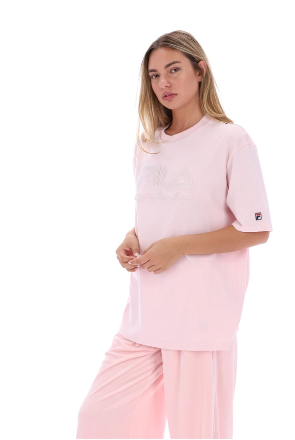 A woman modeling a light pink FILA lounge set, featuring a short-sleeve crewneck t-shirt with a tonal FILA logo on the chest and a small brand patch on the sleeve, paired with matching pink lounge pants, set against a plain white background.