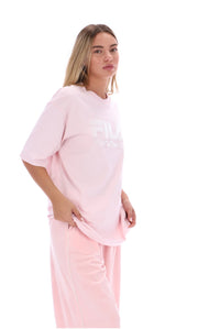 A woman modeling a light pink Fila lounge set, featuring an oversized short-sleeve t-shirt with a white logo and matching wide-leg pants, set against a plain white background.