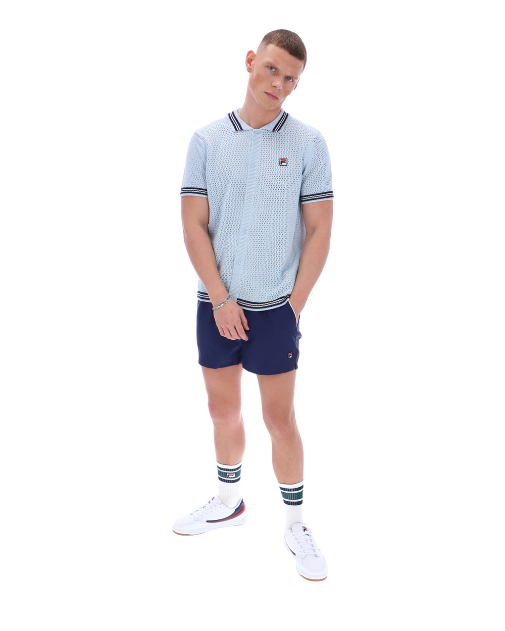 Man wearing a light blue patterned polo shirt with a FILA log on the chest and navy shorts, against a white background.