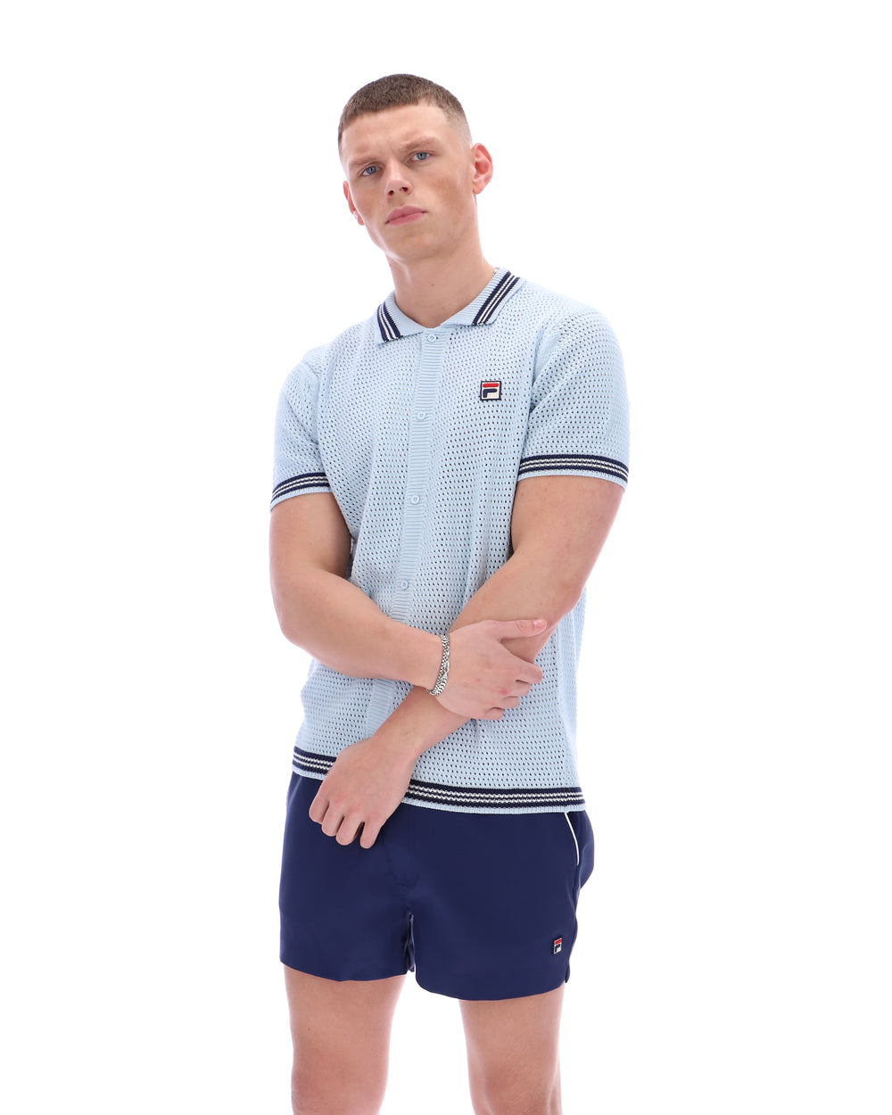Man wearing a light blue patterned polo shirt with a FILA log on the chest and navy shorts, against a white background.