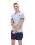 Man wearing a light blue patterned polo shirt with a FILA log on the chest and navy shorts, against a white background.