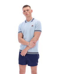 Man wearing a light blue patterned polo shirt with a FILA log on the chest and navy shorts, against a white background.
